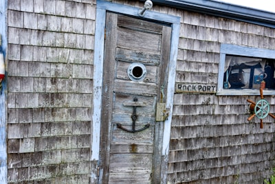 A weathered wooden shack with a rustic appearance, featuring a weathered door with a circular window. An anchor is mounted on the door. There is a sign reading 'ROCKPORT' next to a window with a fishing net and a wooden steering wheel decoration.