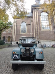 A vintage blue Land Rover with a spare tire mounted on the hood is parked on a cobblestone street in front of a large historic brick building with arched windows. Bicycles are lined up against the building in the background, and tree branches with autumn leaves are overhead.