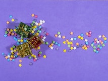Multiple small, colorful star-shaped confetti pieces are scattered across a purple background. Three small gift boxes, decorated with green, red, and gold reflective surfaces and tied with gold string, are placed among the stars.