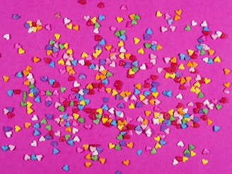 A vibrant scatter of small, colorful heart shapes spread across a pink background, creating a lively and festive pattern.