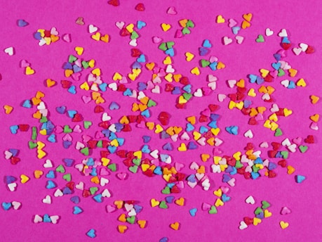 A vibrant scatter of small, colorful heart shapes spread across a pink background, creating a lively and festive pattern.