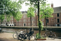 Bicycles are lined up along a canal with a cobblestone path in the foreground. A row of red-brick buildings with white-framed windows and steep roofs lines the opposite side of the canal. Tall green trees provide a lush canopy, and the water in the canal adds a serene atmosphere.