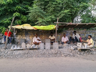 Several men are sitting under a makeshift shelter made of wood and a yellow tarp. They are surrounded by sculptures and objects made of stone, which seem to be handmade. The men are seated on a patch of rough gravel next to a wall, with dense green foliage in the background. The atmosphere suggests a roadside market or workshop, possibly in a rural or semi-urban setting.