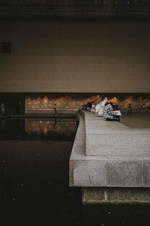 Several people are sitting on the edge of a wide, concrete platform beside a reflecting pool inside a modern, minimalist building with high ceilings. The scene is dimly lit with warm, ambient light reflecting off the water's surface.