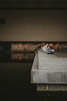 Several people are sitting on the edge of a wide, concrete platform beside a reflecting pool inside a modern, minimalist building with high ceilings. The scene is dimly lit with warm, ambient light reflecting off the water's surface.