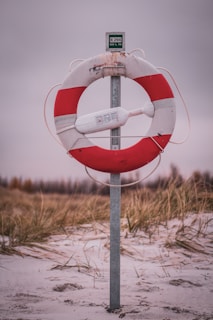 A life preserver mounted on a post, situated on a sandy beach with tall grasses in the background. The life preserver is red and white, with a safety device attached.