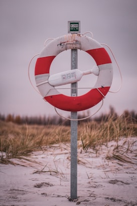 A life preserver mounted on a post, situated on a sandy beach with tall grasses in the background. The life preserver is red and white, with a safety device attached.