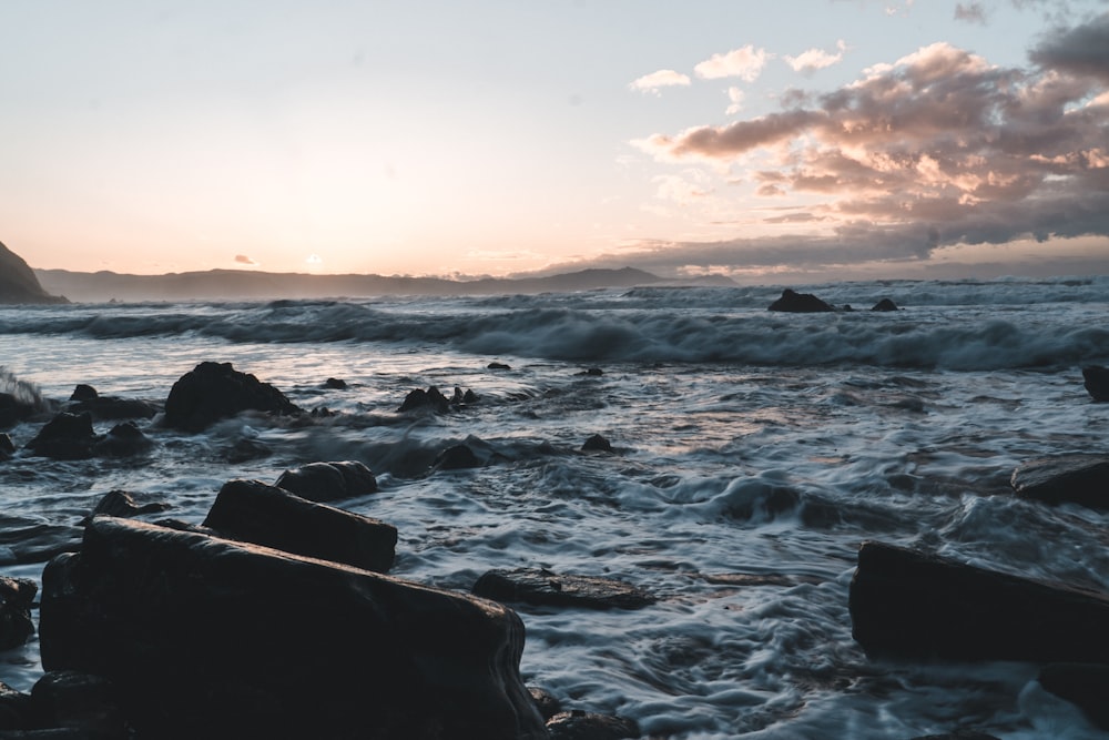 the sun is setting over the ocean with rocks in the foreground