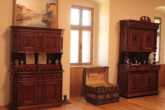 An interior room with wooden furniture, including two ornate wooden cabinets and an open antique trunk. The left cabinet has several drawers and some decorative items on its shelves, with a painting hanging above it. The right cabinet is similarly ornate with dishes and decorative objects on its shelves. A set of windows is situated between the two cabinets, allowing natural light to fill the room.