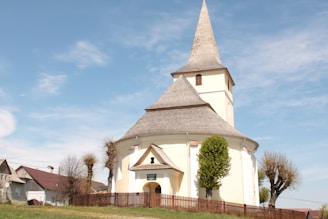 A quaint, round church with a tall, pointed steeple stands surrounded by a simple wooden fence. The church is painted in a pale shade and has a distinctive architectural design. There are a few small trees and other buildings in the background under a clear blue sky, creating a serene and peaceful rural setting.