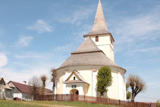 A quaint, round church with a tall, pointed steeple stands surrounded by a simple wooden fence. The church is painted in a pale shade and has a distinctive architectural design. There are a few small trees and other buildings in the background under a clear blue sky, creating a serene and peaceful rural setting.