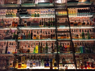 A wall displaying a large collection of liquor bottles arranged on glass shelves with a metal ladder leaning against them. Soft lighting highlights the various shapes and colors of the bottles, set against a background of red brick.