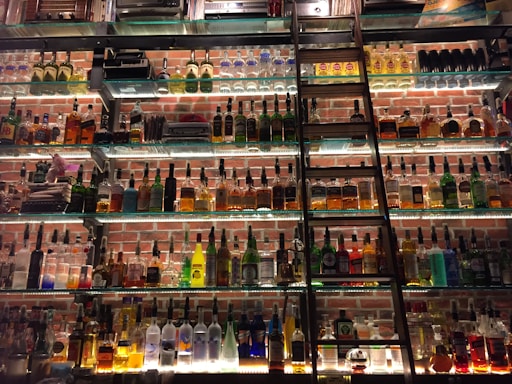 A wall displaying a large collection of liquor bottles arranged on glass shelves with a metal ladder leaning against them. Soft lighting highlights the various shapes and colors of the bottles, set against a background of red brick.