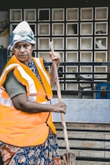 An individual wearing an orange safety vest and a makeshift head covering, holding a broom, with a background of a tiled wall and some visible furniture.