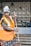 An individual wearing an orange safety vest and a makeshift head covering, holding a broom, with a background of a tiled wall and some visible furniture.