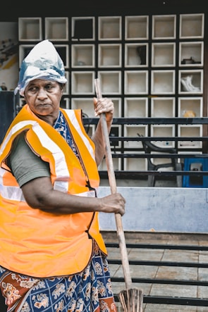 An individual wearing an orange safety vest and a makeshift head covering, holding a broom, with a background of a tiled wall and some visible furniture.