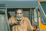 A man with gray hair and a beard is sitting in a rickshaw, holding a phone in his hand. He is wearing a brown shirt and looking directly at the camera with a warm, friendly expression. There are droplets of water on the rickshaw, indicating it has rained recently.