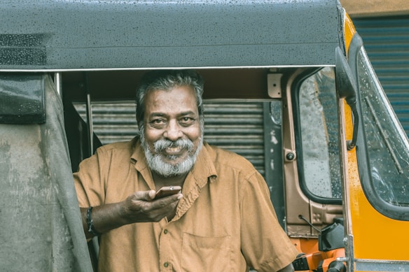 A man with gray hair and a beard is sitting in a rickshaw, holding a phone in his hand. He is wearing a brown shirt and looking directly at the camera with a warm, friendly expression. There are droplets of water on the rickshaw, indicating it has rained recently.