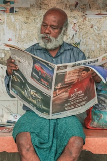 An older man with a white beard and bald head is sitting on a bench, deeply absorbed in reading a newspaper. He is wearing a blue shirt and a patterned wrap or lungi. The background features a worn wall with various pieces of paper and posters partially peeling off. Beside him, a colorful bag rests on the bench.