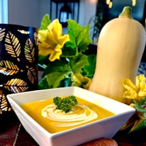 A creamy bowl of butternut squash soup is garnished with a swirl of cream and a sprig of parsley, presented in a white square bowl. In the background, a whole butternut squash and lush green leaves with yellow flowers are visible. A dark candle holder with a leaf pattern and soft ambient lighting gives the setting a warm, cozy atmosphere.