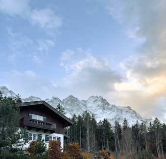 A quaint alpine house situated at the base of snow-capped mountains. The house features a wooden upper level and a white lower level, surrounded by tall evergreen trees and autumn-colored foliage. Above, the sky displays a blend of blue and pastel-colored clouds, casting a serene and peaceful atmosphere.