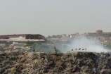 A large pile of waste with visible smoke rising, set against a backdrop of urban buildings. A person is seen on or near a bicycle on the pile, and several birds are perched on the rubbish.