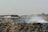 A large pile of waste with visible smoke rising, set against a backdrop of urban buildings. A person is seen on or near a bicycle on the pile, and several birds are perched on the rubbish.