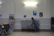 A person wearing a gray coat and blue gloves is cleaning a table in a room with a tiled floor and cream-colored walls. There are several posters on the wall and a light fixture above. To the left, there are stacked chairs and some tables.