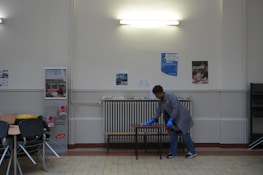 A person wearing a gray coat and blue gloves is cleaning a table in a room with a tiled floor and cream-colored walls. There are several posters on the wall and a light fixture above. To the left, there are stacked chairs and some tables.