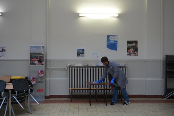 A person wearing a gray coat and blue gloves is cleaning a table in a room with a tiled floor and cream-colored walls. There are several posters on the wall and a light fixture above. To the left, there are stacked chairs and some tables.
