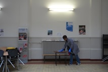 A person wearing a gray coat and blue gloves is cleaning a table in a room with a tiled floor and cream-colored walls. There are several posters on the wall and a light fixture above. To the left, there are stacked chairs and some tables.