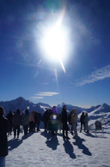 Energetic group of snowscoot riders celebrating on a snowy mountain peak under a bright blue sky.