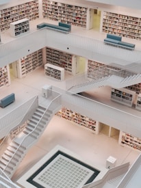 A spacious, modern library with multiple levels lined with bookshelves. Clean, minimalist design with white floors and railings. Several open staircases connect the different levels, and the layout is symmetrical and geometric. Comfortable seating areas are sparsely placed along the edges.