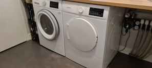 Two white front-loading laundry machines are placed side by side beneath a wooden countertop. The left machine appears to be a washing machine while the right one is a dryer. They are situated in a utility room with a grey floor and some plumbing fixtures visible to the right.