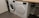 Two white front-loading laundry machines are placed side by side beneath a wooden countertop. The left machine appears to be a washing machine while the right one is a dryer. They are situated in a utility room with a grey floor and some plumbing fixtures visible to the right.