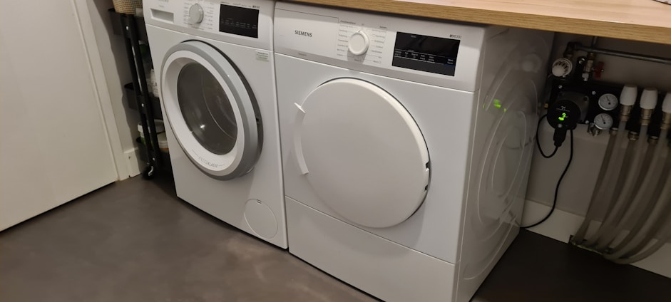 Two white front-loading laundry machines are placed side by side beneath a wooden countertop. The left machine appears to be a washing machine while the right one is a dryer. They are situated in a utility room with a grey floor and some plumbing fixtures visible to the right.
