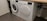 Two white front-loading laundry machines are placed side by side beneath a wooden countertop. The left machine appears to be a washing machine while the right one is a dryer. They are situated in a utility room with a grey floor and some plumbing fixtures visible to the right.