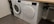 Two white front-loading laundry machines are placed side by side beneath a wooden countertop. The left machine appears to be a washing machine while the right one is a dryer. They are situated in a utility room with a grey floor and some plumbing fixtures visible to the right.
