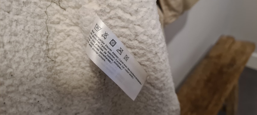 A close-up of a clothing care label attached to a cream-colored, textured fabric. Care symbols and instructions are printed on the label.