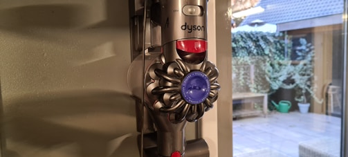 A Dyson vacuum cleaner is mounted on a wall next to a window. Outside the window, there is a patio with plants and a green watering can. The room has a soft, natural light coming in.
