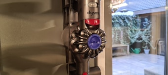 A Dyson vacuum cleaner is mounted on a wall next to a window. Outside the window, there is a patio with plants and a green watering can. The room has a soft, natural light coming in.