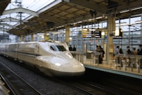 A sleek, modern high-speed train is captured arriving at a station. The platform is bustling with passengers, some of whom are glancing at overhead electronic boards displaying information. The station has a contemporary design with metal and glass structures.