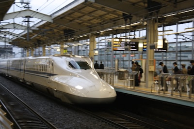 A sleek, modern high-speed train is captured arriving at a station. The platform is bustling with passengers, some of whom are glancing at overhead electronic boards displaying information. The station has a contemporary design with metal and glass structures.