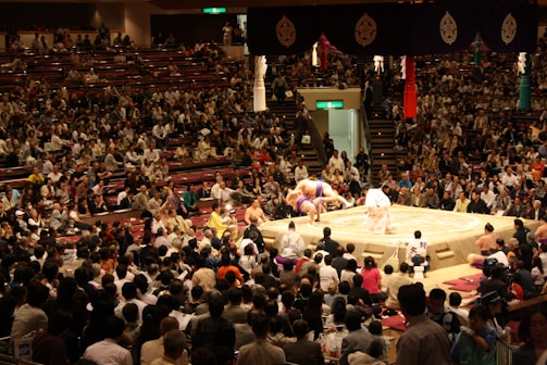 A large crowd fills a sumo wrestling arena, engaging in watching a sumo match on a prominent elevated ring in the center. The wrestlers and a referee are focused on the bout, surrounded by many spectators seated in rows. Traditional decorations hang above and around the ring.