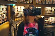 A person standing in a well-lit library, wearing a virtual reality headset and headphones. The individual has long hair and is dressed in a colorful T-shirt depicting an astronaut. The background features numerous bookshelves filled with books.
