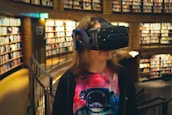 A person standing in a well-lit library, wearing a virtual reality headset and headphones. The individual has long hair and is dressed in a colorful T-shirt depicting an astronaut. The background features numerous bookshelves filled with books.