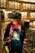 A person wearing a virtual reality headset standing in a library. They hold VR controllers in both hands and wear a colorful shirt featuring an astronaut graphic. The shelves are lined with numerous books.