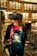 A person wearing a virtual reality headset standing in a library. They hold VR controllers in both hands and wear a colorful shirt featuring an astronaut graphic. The shelves are lined with numerous books.