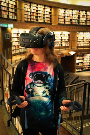 A person wearing a virtual reality headset standing in a library. They hold VR controllers in both hands and wear a colorful shirt featuring an astronaut graphic. The shelves are lined with numerous books.