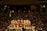 Several sumo wrestlers are gathered in a circle on a sumo wrestling ring surrounded by a large crowd of spectators in a dimly lit arena. The wrestlers are wearing traditional mawashi belts of various colors, and the scene is lit from above, creating a focused and intense atmosphere.